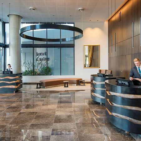 Pullman Brisbane Airport Hotel Luaran gambar The image shows a modern hotel lobby. There are two reception desks made of dark-colored materials, and staff members are attending to guests at these desks. The lobby features large windows that allow natural light to fill the space, and there are p